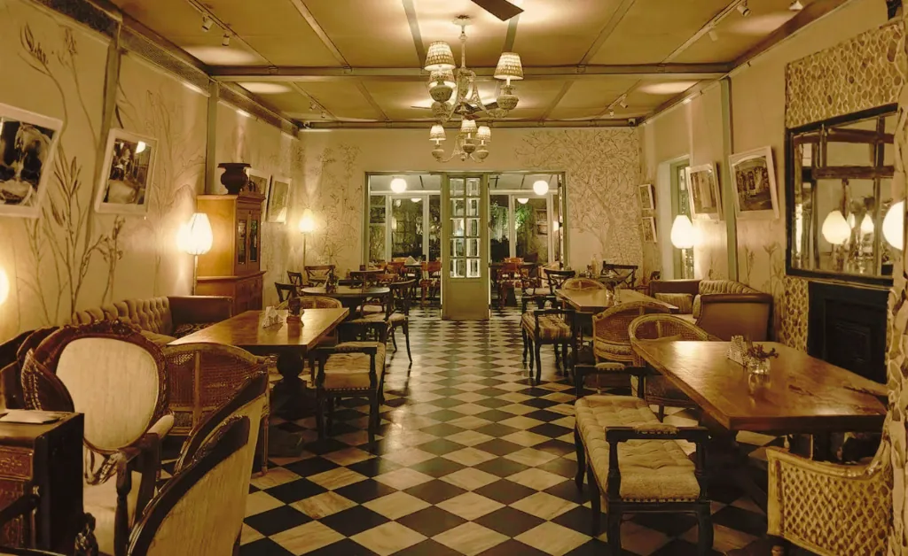 Classic cafe interior with checkered floor, vintage wooden tables, and wicker chairs. Soft lighting from lamps and chandeliers creates a cozy ambiance.