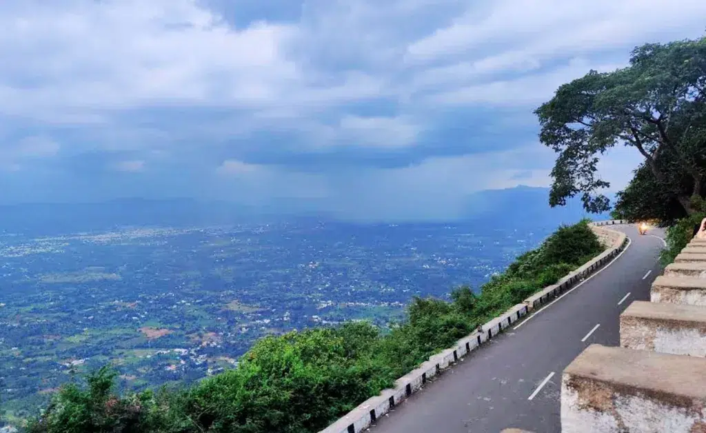 A serene mountain road curves along a lush, green hillside under an expansive, cloudy sky. The valley below is dotted with greenery, conveying tranquility.